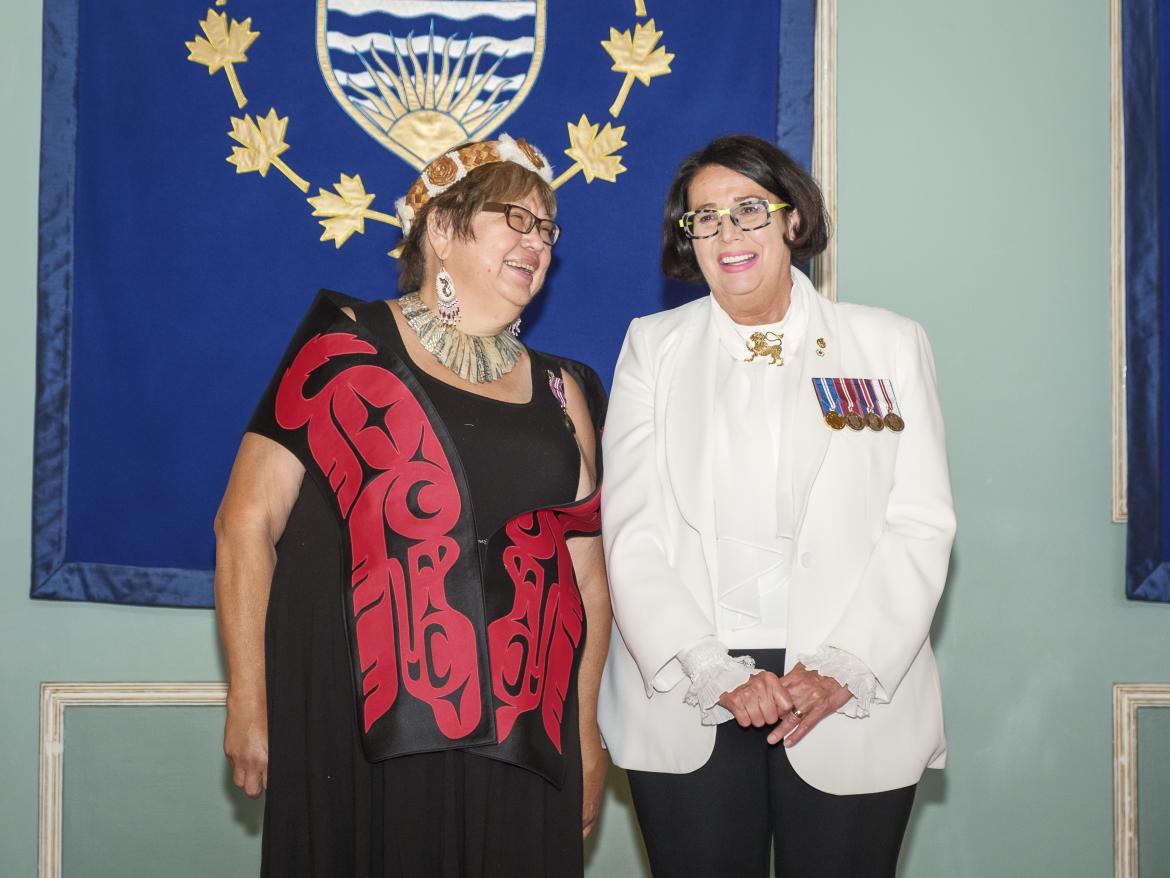 Two people pose in front of the BC coat of arms