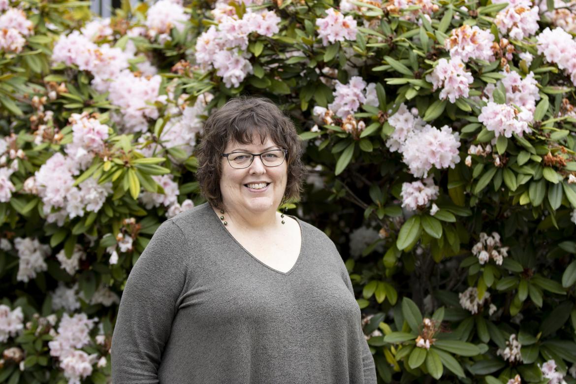 Terri Doughty, a VIU English Professor, smiles while standing in front of a rhododendron plant.