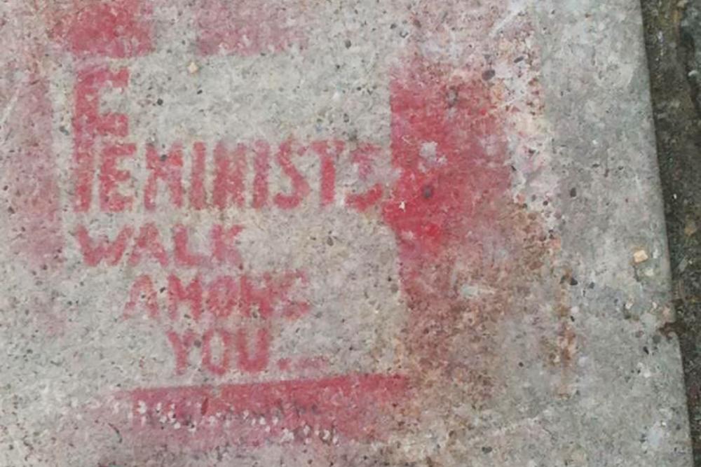 A light grey concrete sidewalk is spray painted with the phrase "Feminists Walk Among You."