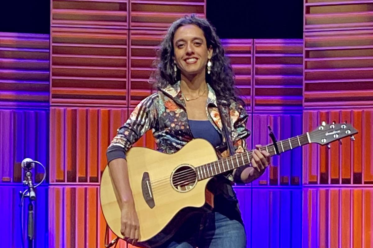 Sonnet L’Abbé sits on a stool on the Port Theatre's stage in front of a multi-coloured pink, blue and purple lighted background.